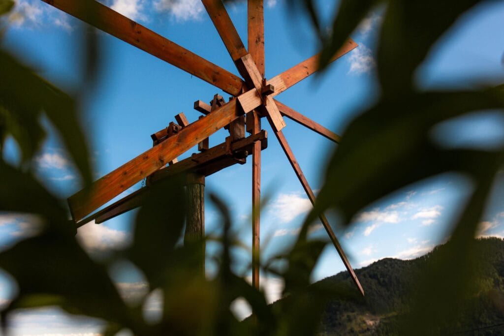 Alter Windmühlenflügel vor blauem Himmel und Wolken, umgeben von grünen Blättern.